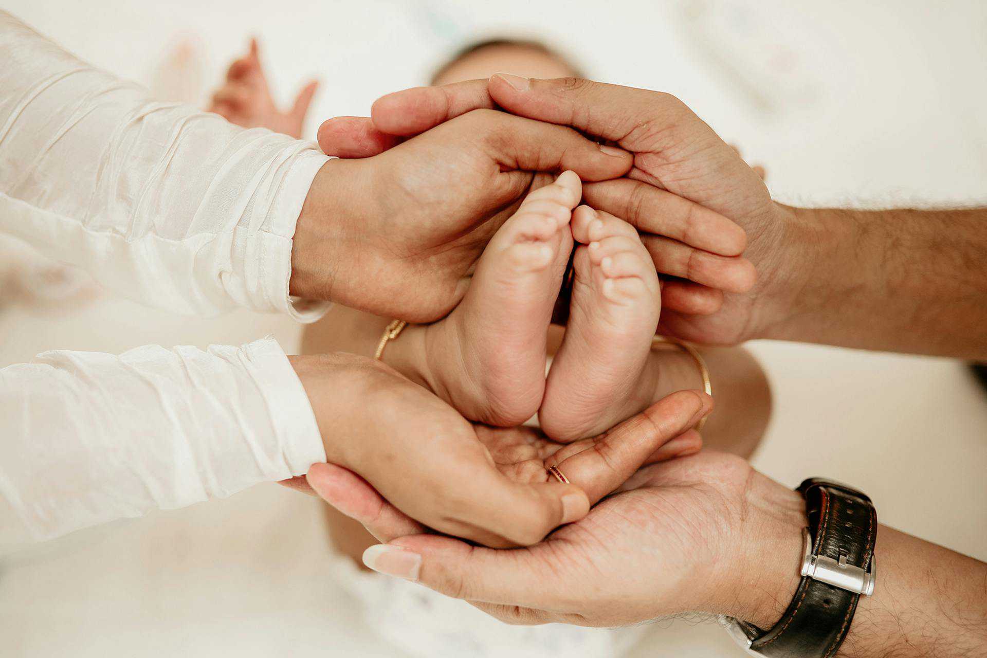 A baby's feet are cupped by its mother's hands, which are in turn cupped by the father's hands.