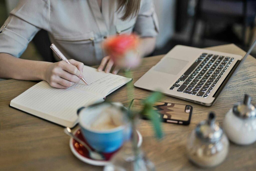 Someone researching at their desk.  A laptop is open and a woman is writing notes in her notebook.  A coffee is next to her on the table.