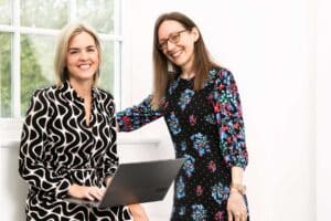 Photo of Clara and Shannon from Downton Mortgages & Financial Services smiling at the camera in front of a window. Shannon has a laptop open in front of her.