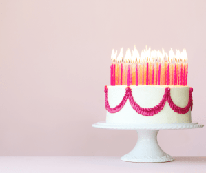 A birthday cake with lots and lots of candles sits on a cake stand with a pink background behind it.