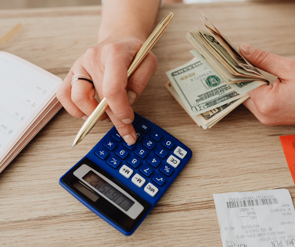 A woman types numbers into a blue calculator. She has a pencil in her hand, making notes. She also holds a wad of cash in her other hand.